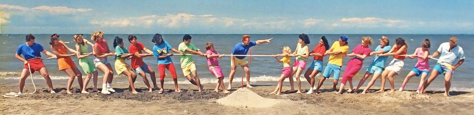 Two groups of people in brightly colored clothes on a beach playing tug of war with each other.