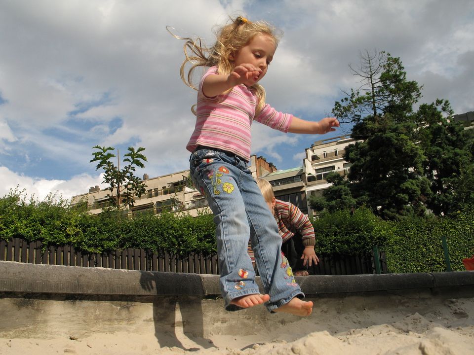 Close up of a young (4-5 year old) blonde child jumping into a sandbox. Trees and a building are in the background.